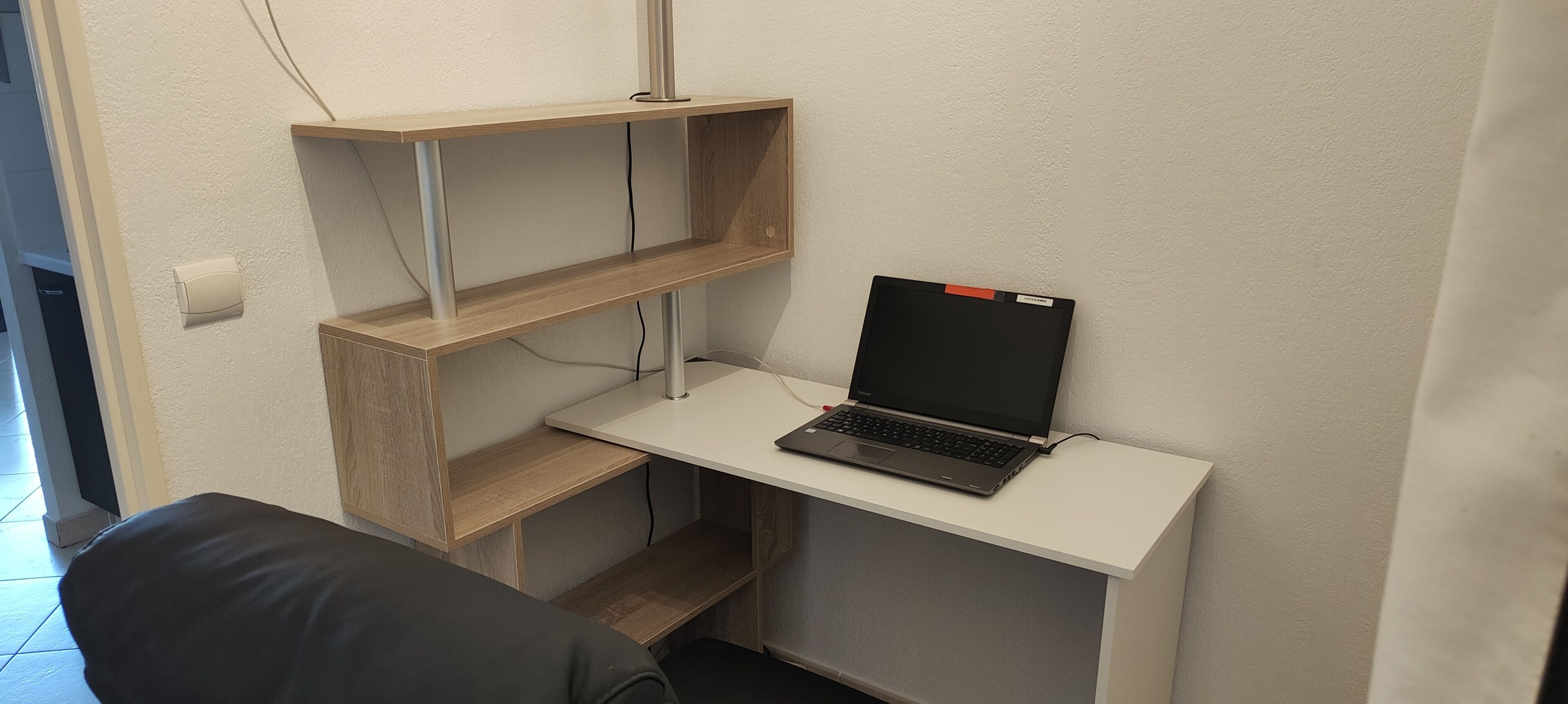 A compact workspace with a laptop on a white desk, wooden shelves, and a black chair.