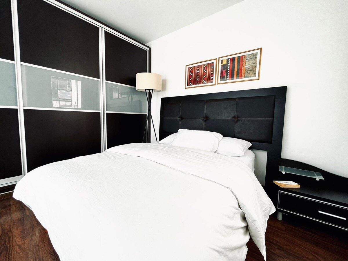 Une chambre moderne avec un grand lit, une tête de lit sombre, une table de chevet avec une lampe et une armoire aux portes miroir.
