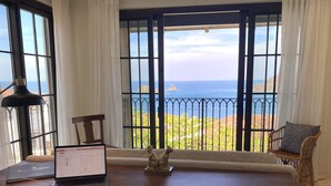 Ein Zimmer mit Schreibtisch aus Holz, einem Laptop, Papieren und einer dekorativen Hundefigur. Ein Fenster mit Meerblick und ein Balkon mit einem Stuhl sind zu sehen.