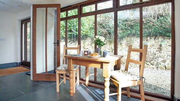 Een kamer met een houten tafel, twee stoelen, een vaas met bloemen en uitzicht op groen door grote ramen.