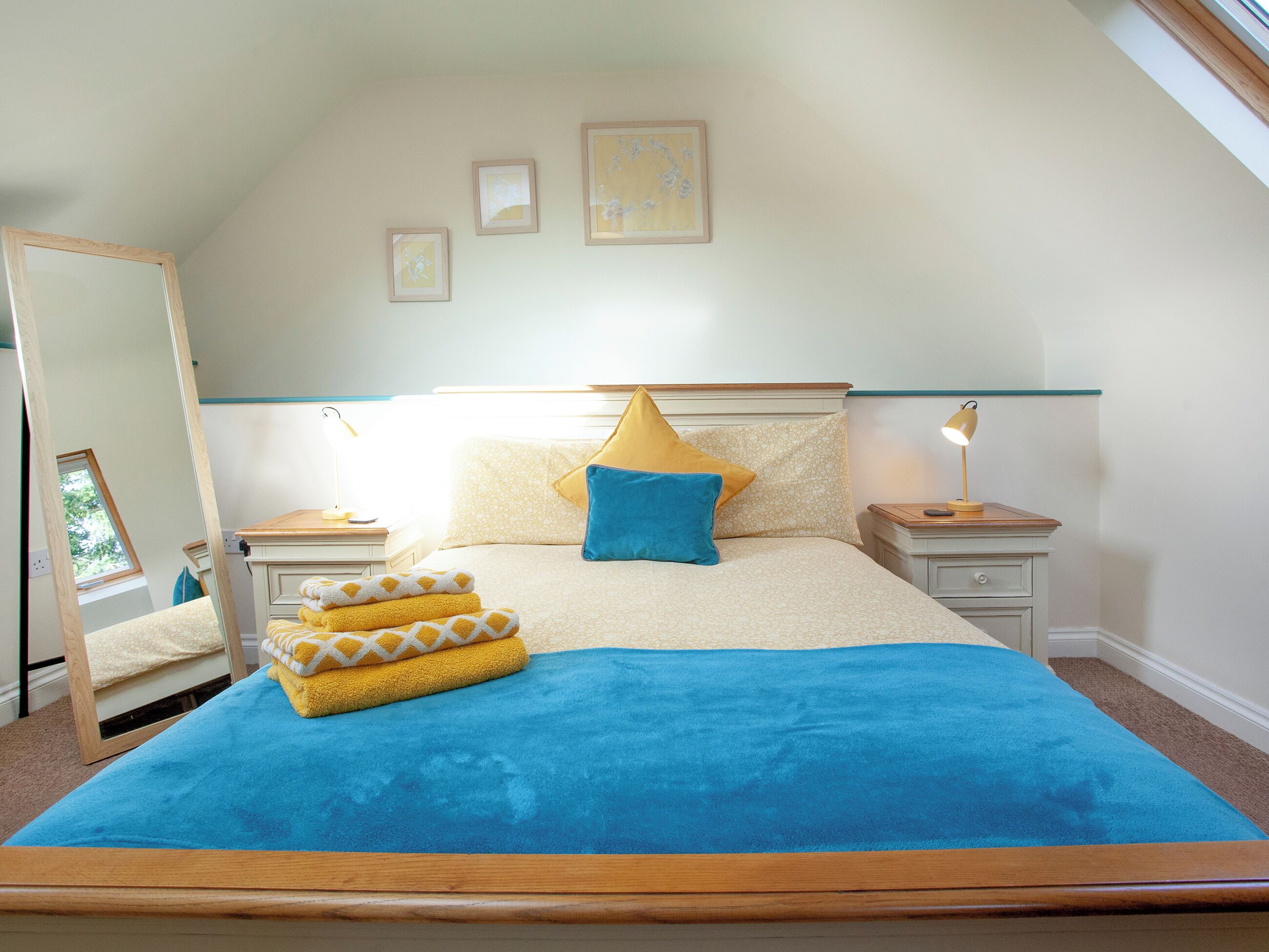 A bedroom with a bed, two bedside tables, a nightstand, a mirror, and three framed pictures on the wall.
