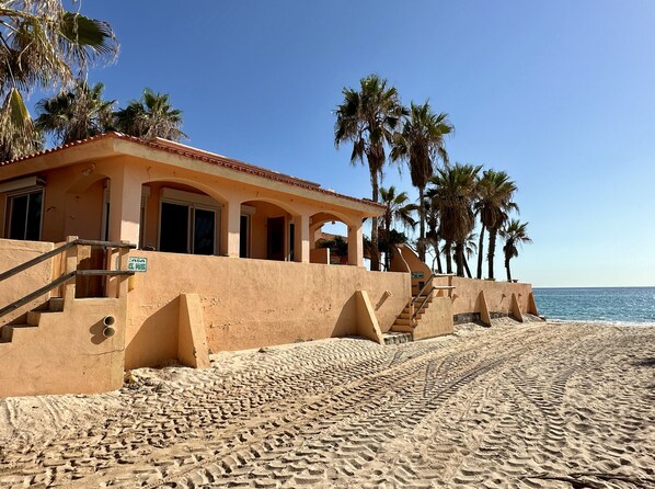 Ein direkt am Strand gelegenes Anwesen mit Putzfassade, einer Treppe zu einer überdachten Terrasse und Palmen im Hintergrund.