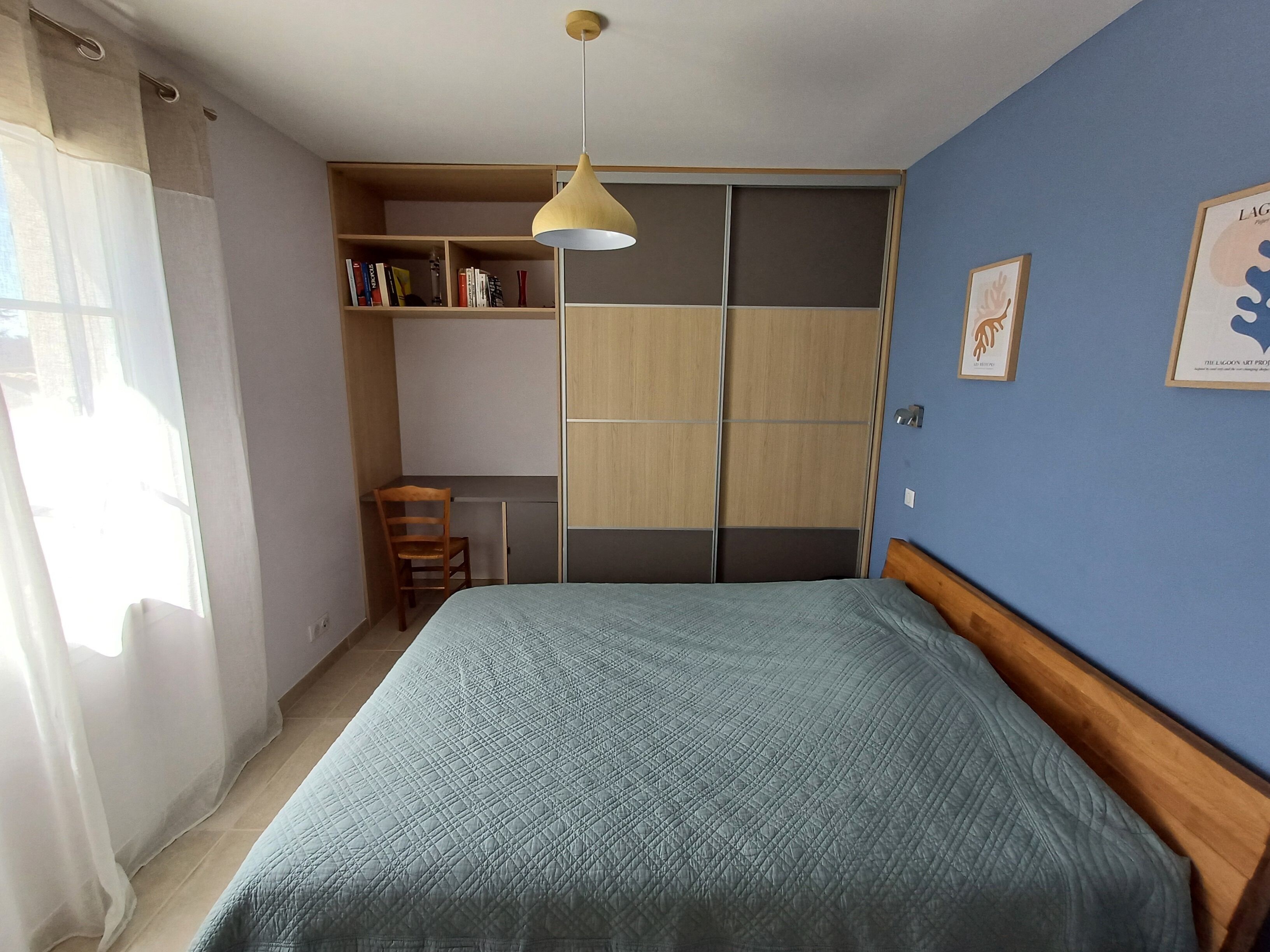 A bedroom with a bed, a wooden wardrobe, a chair, and a shelf with books.