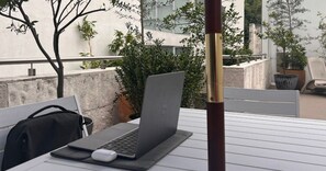 A laptop on a table with a mouse, a black bag, and a chair in an outdoor setting with plants and a building in the background.