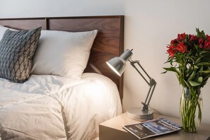 Un lit bien fait, avec du linge de lit blanc, un coussin à motifs, une lampe de chevet, un magazine et un vase rempli de fleurs.