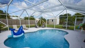 A covered swimming pool with a blue inflatable shark, lounge chairs, and a wooden fence in the background. - Luxurious Pool Cottage Sleep 2 (Port St. Lucie)