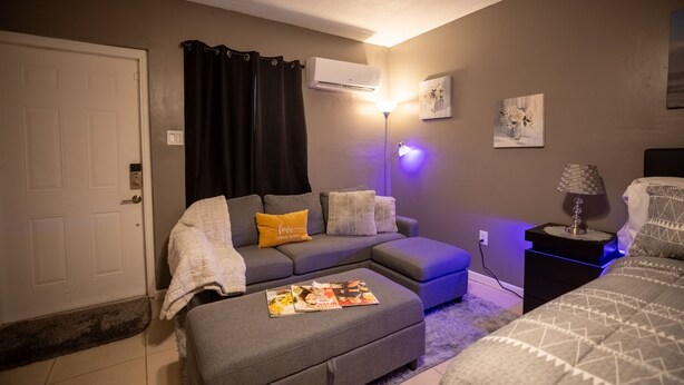 A living room with a grey sofa, a small ottoman with magazines, a lamp, and a painting on the wall. - Luxurious Pool Cottage Sleep 2 (Port St. Lucie)