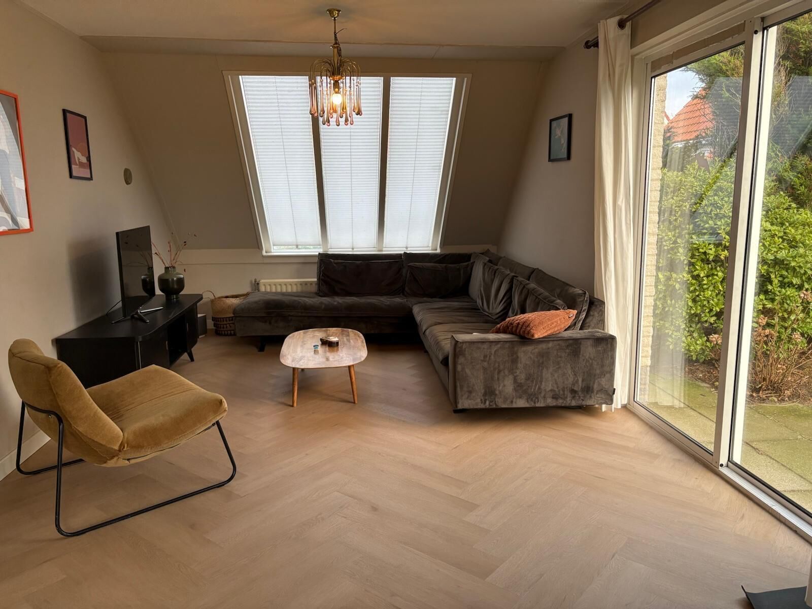 A modern living room with a sectional sofa, a small round coffee table, and a single armchair. There is a large window with blinds, a ceiling light fixture, and a view of greenery outside.