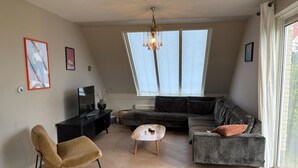 A modern living room with a sofa, armchair, coffee table, and a TV on a black stand. There is a large window with blinds and a chandelier. - Luxe Freistehendes Familie Ferien-haus mit Sauna (Scharendijke)