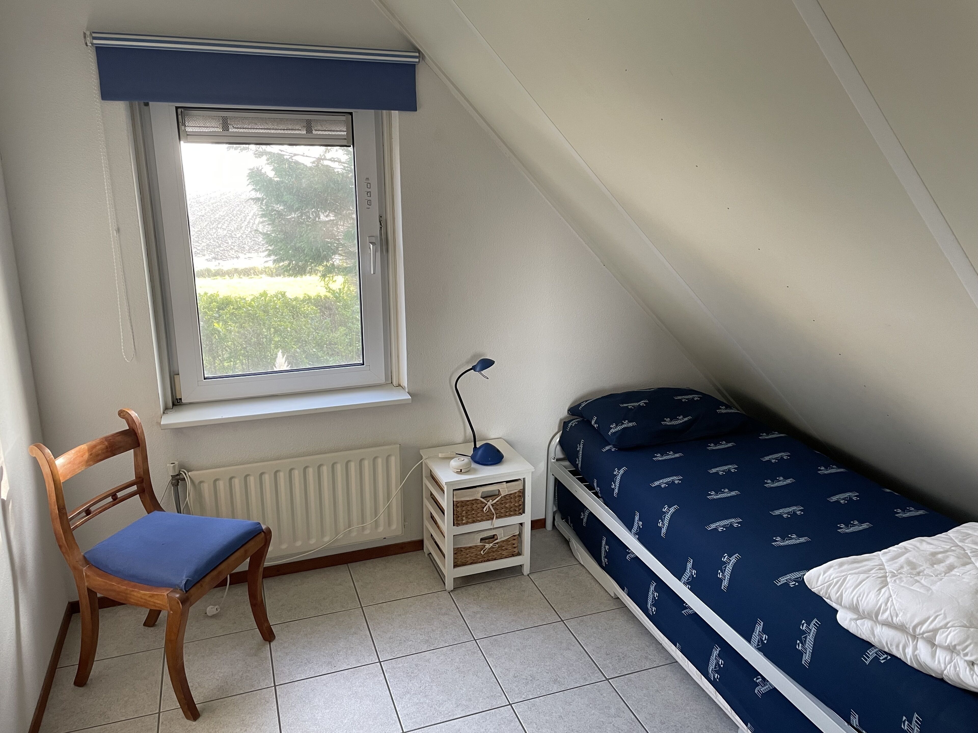 A small, well-lit room with a sloped ceiling, a single bed with a patterned duvet, a wooden chair with a cushion, a desk with a lamp, and a window with a view of greenery outside.