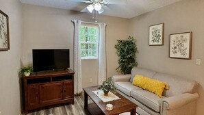 A living room with a sofa, a coffee table, a TV on a stand, a ceiling fan, and a window with curtains.