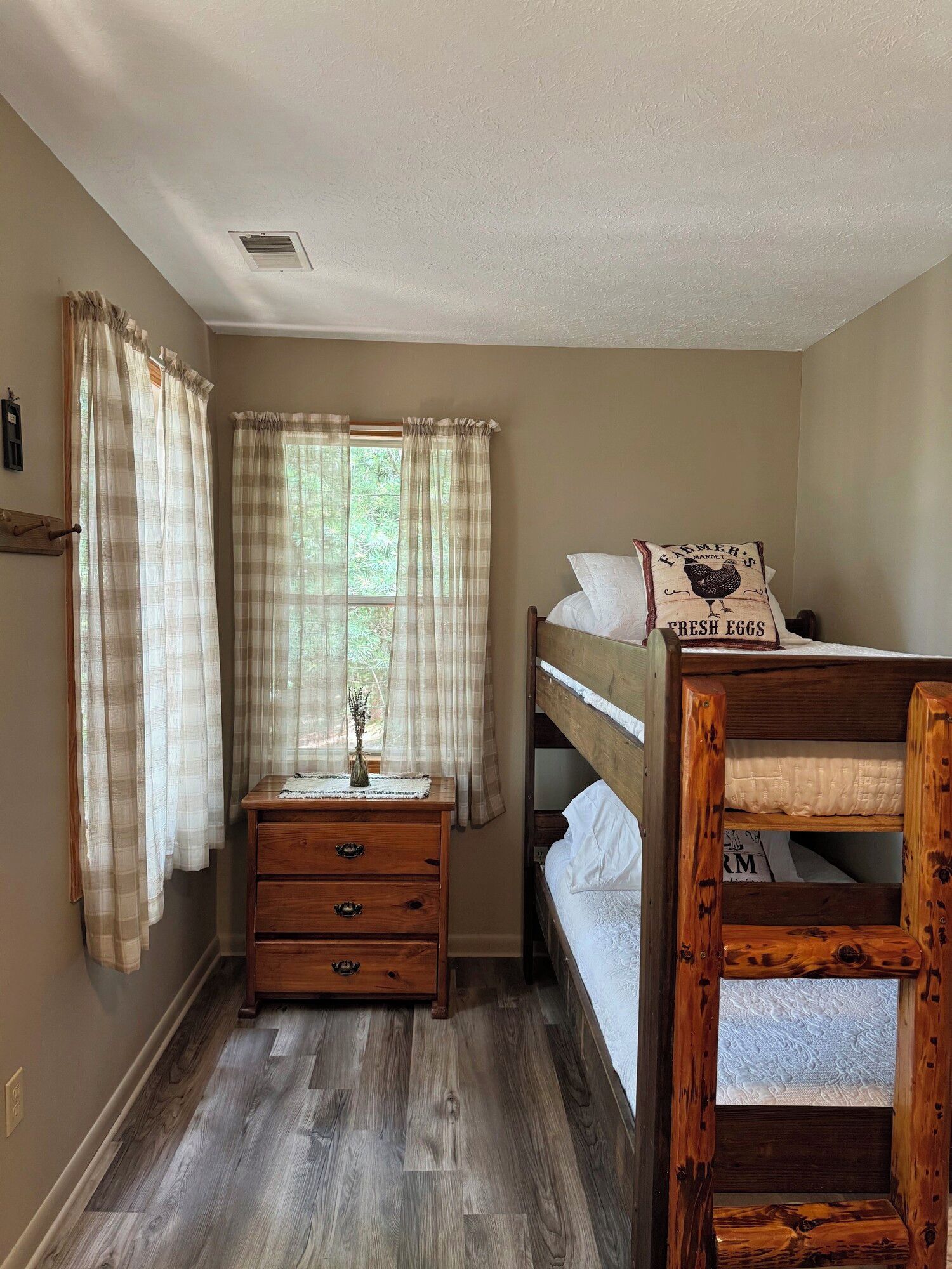 Une chambre avec un lit superposé en bois, une commode en bois avec des tiroirs, une fenêtre avec des rideaux légers et une petite table de chevet avec un vase.