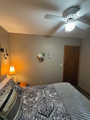 A bedroom with a bed, a nightstand, a lamp, a ceiling fan, and a door.
