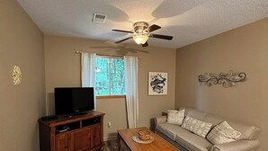 Una sala de estar con ventilador de techo, televisor sobre un mueble de madera, sofá, mesa de centro y ventana con cortinas.