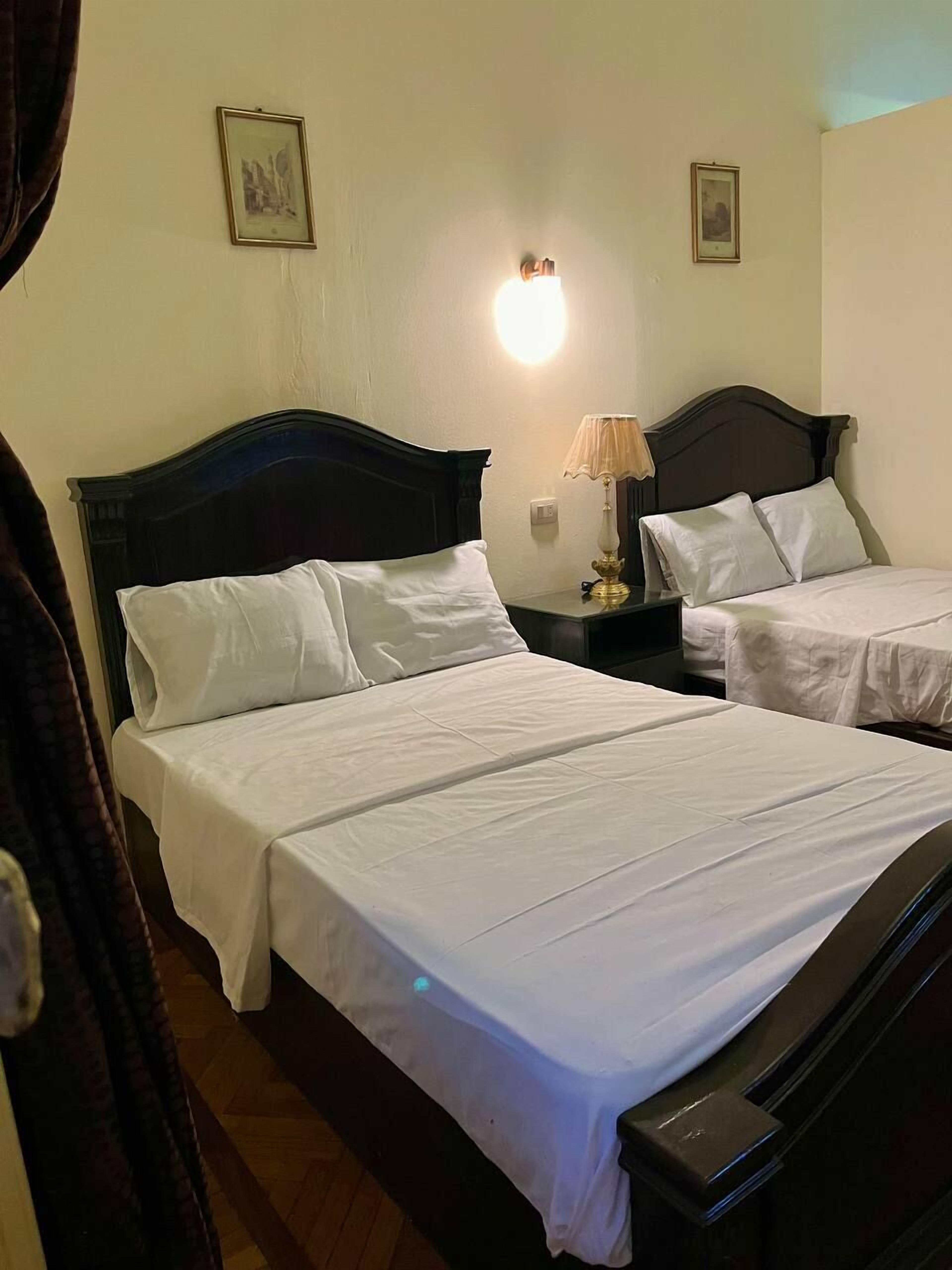 A hotel room with two beds, a nightstand with a lamp, and a wall-mounted light.