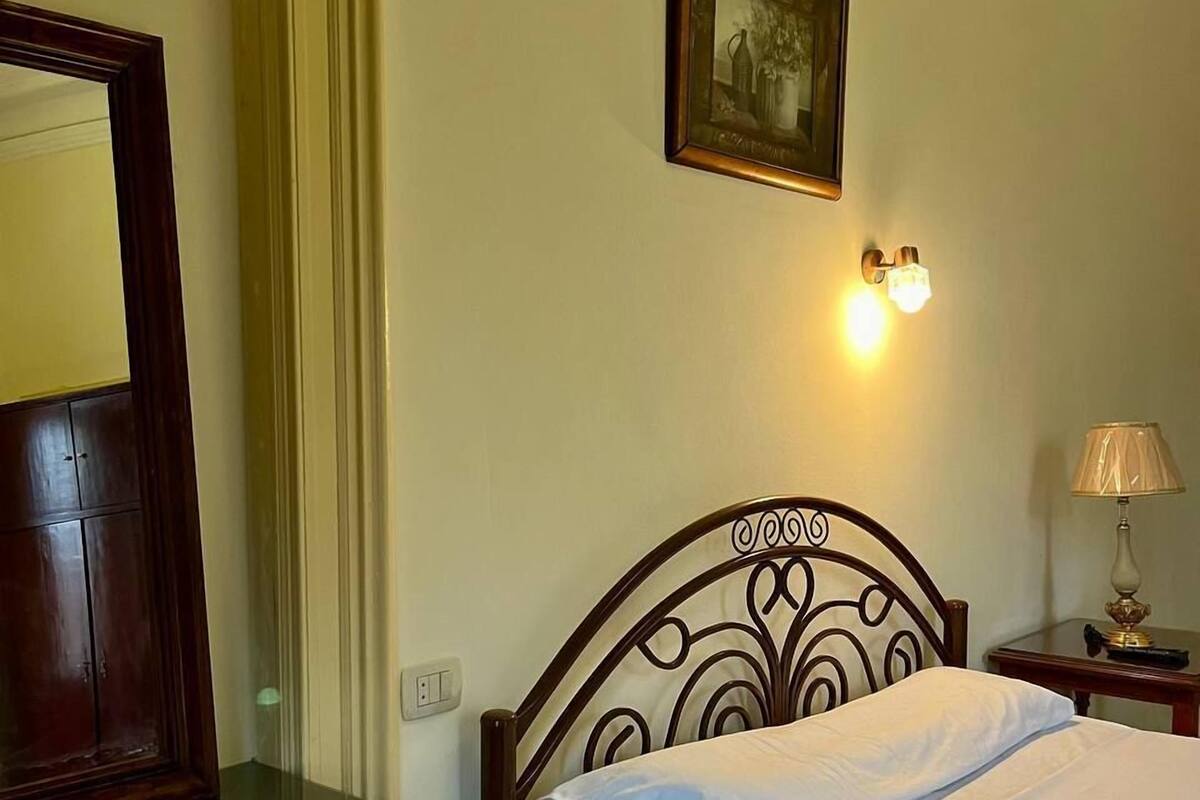 A neatly made bed with white linens, a wooden headboard with a decorative design, a bedside table with a lamp, and a framed picture on the wall.