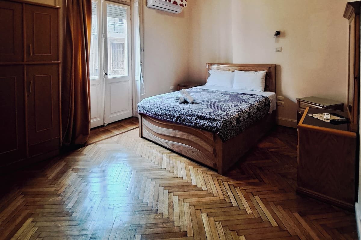 A bedroom with a wooden herringbone floor, a bed with a patterned bedspread, a nightstand with a lamp, and an air conditioning unit on the wall.