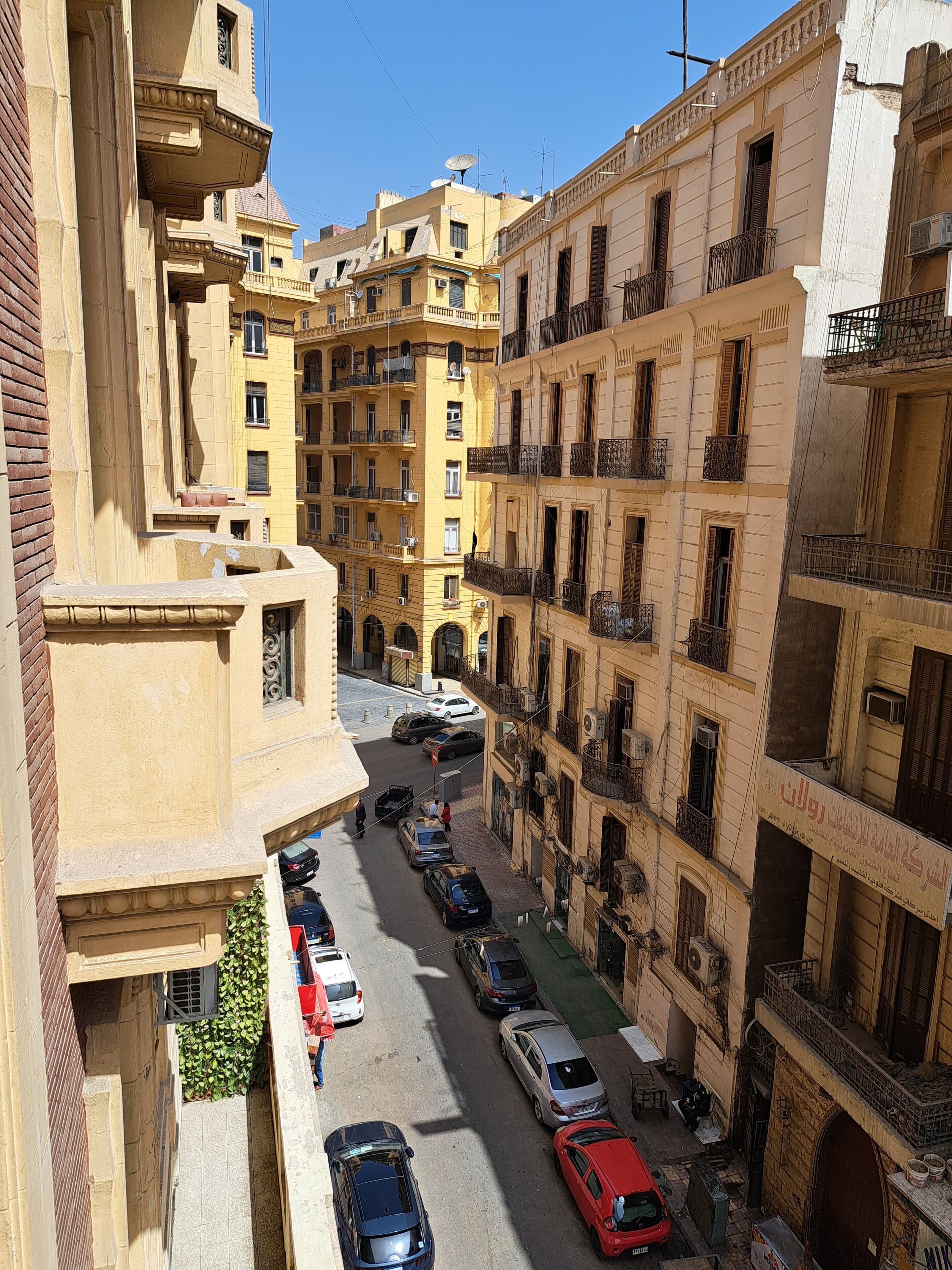 Una calle estrecha bordeada por edificios de varios pisos con balcones, coches aparcados y un cielo azul despejado.