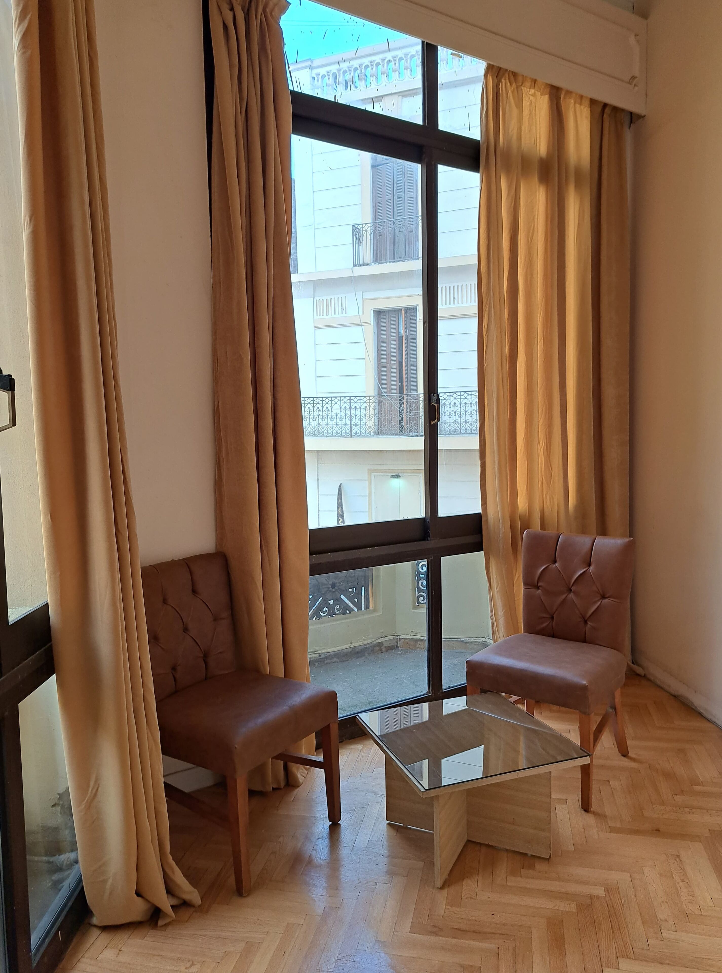 A room with two brown upholstered chairs, a glass coffee table, and large windows with beige curtains.