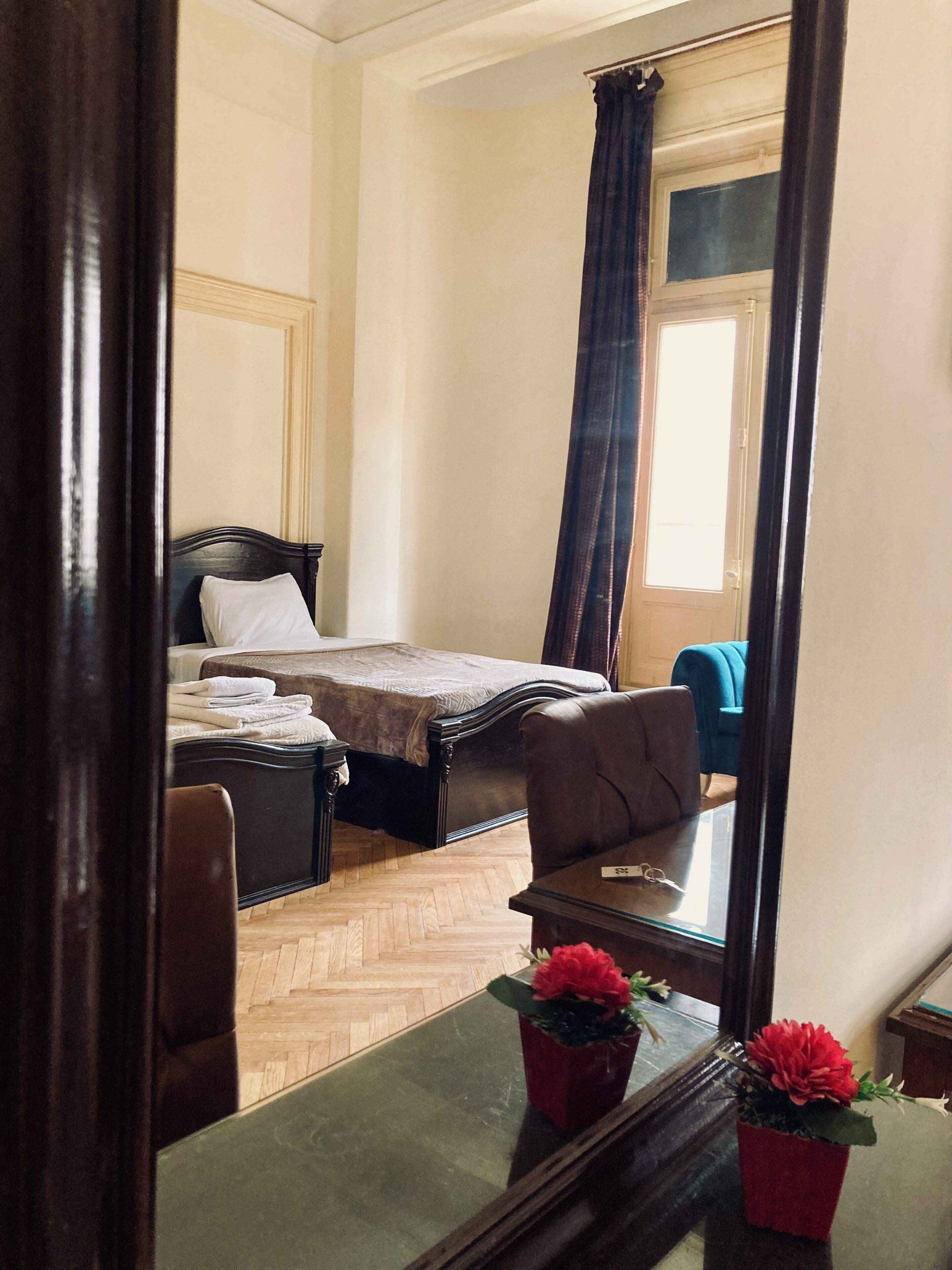 Un dormitorio con una cama, una silla, una mesa con un florero y un espejo que refleja la habitación.