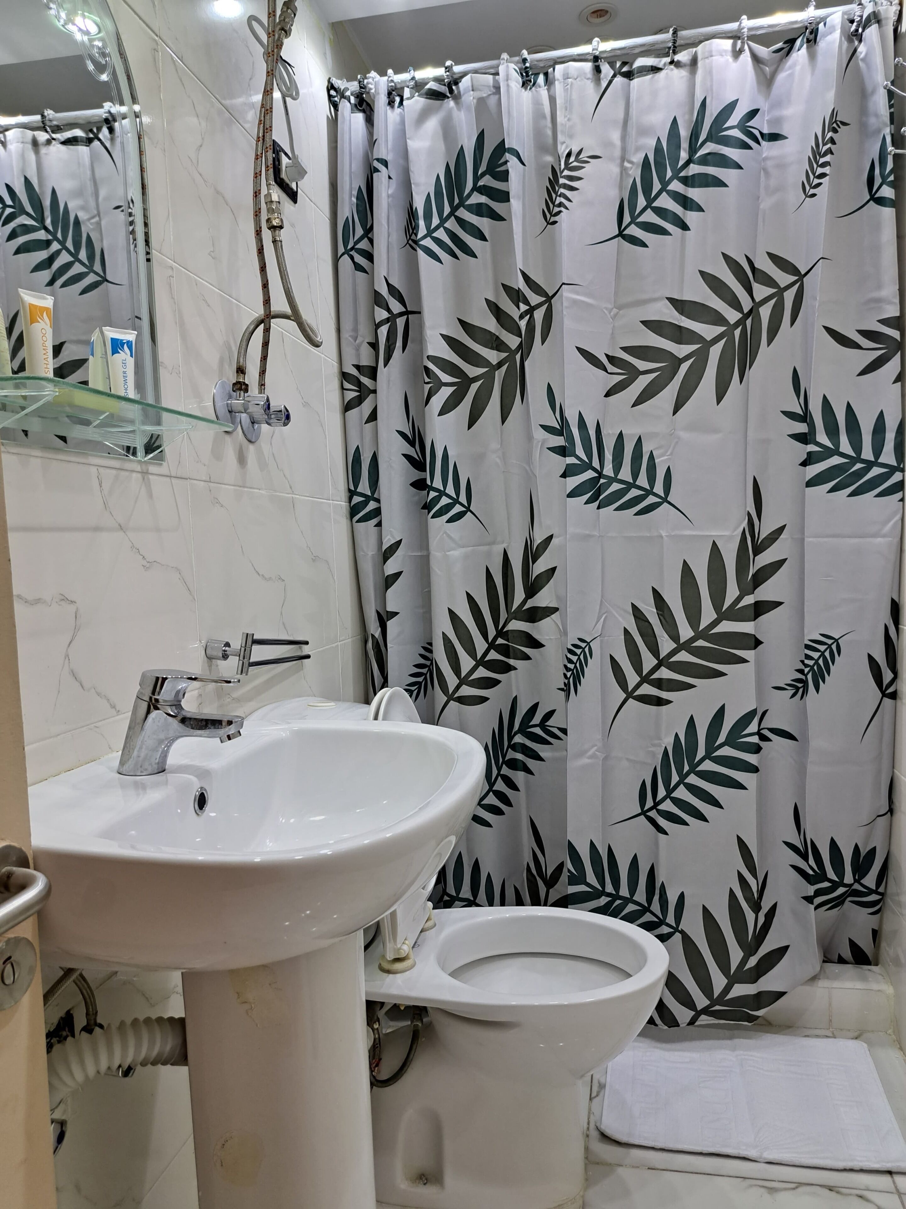 Un baño con lavamanos, inodoro y cortina de ducha blancos, con estampado de hojas.