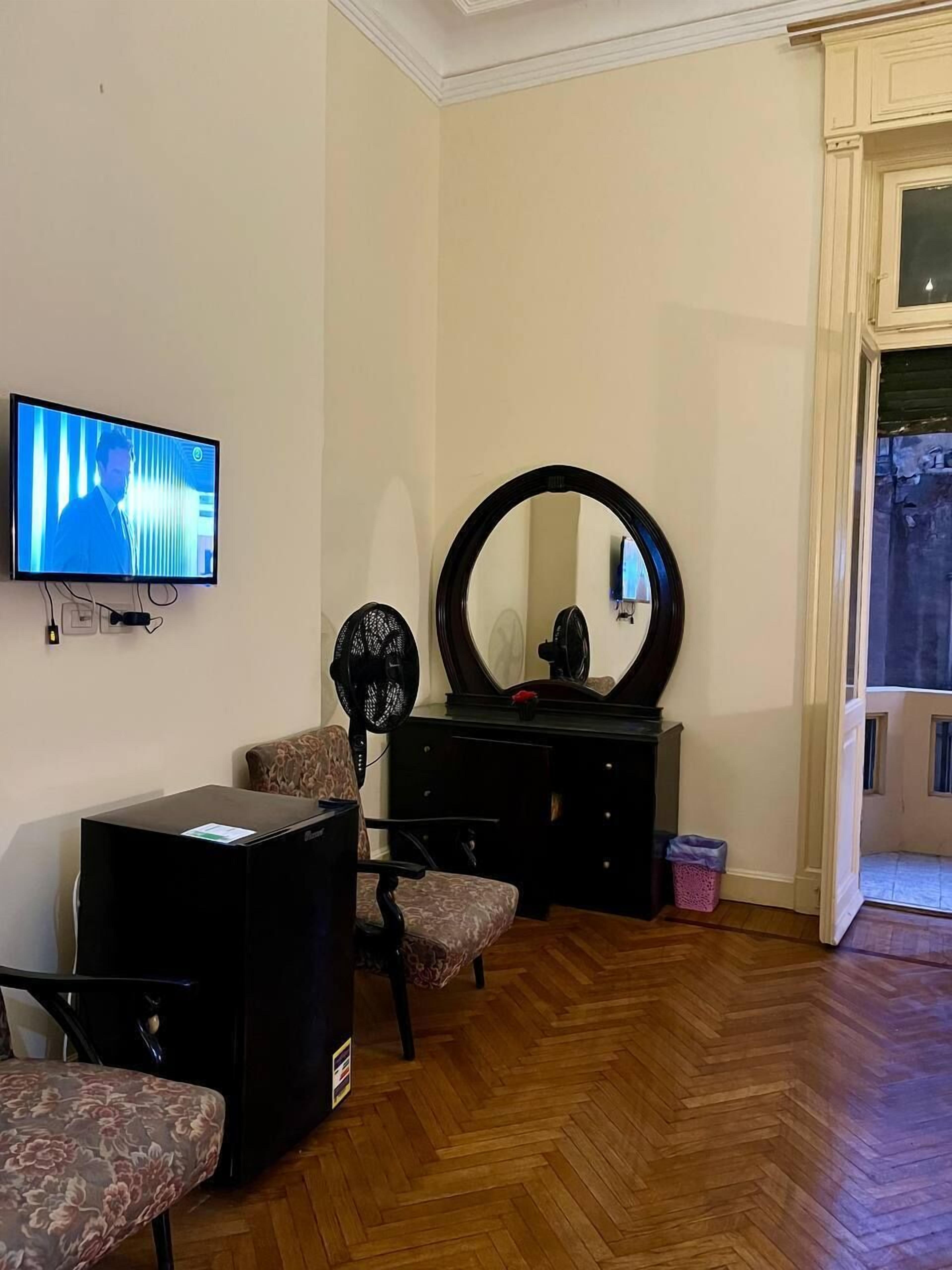 Una habitación con televisor, un espejo grande, una cómoda, una silla y un cubo de basura pequeño.