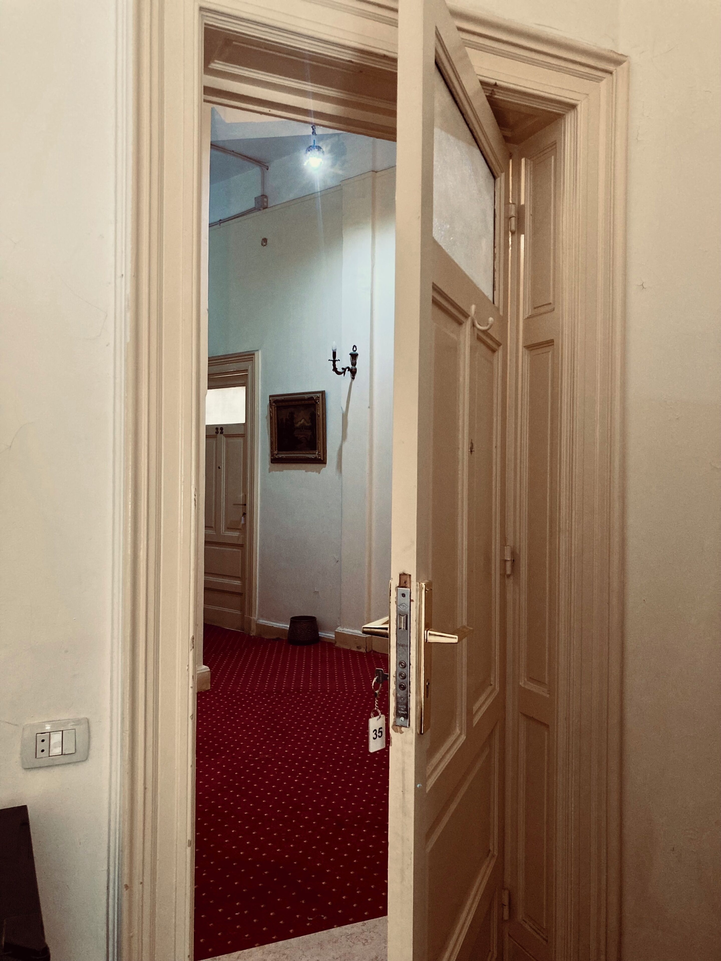 A hallway with a red carpet, a door with a number plate, and a wall-mounted light fixture.