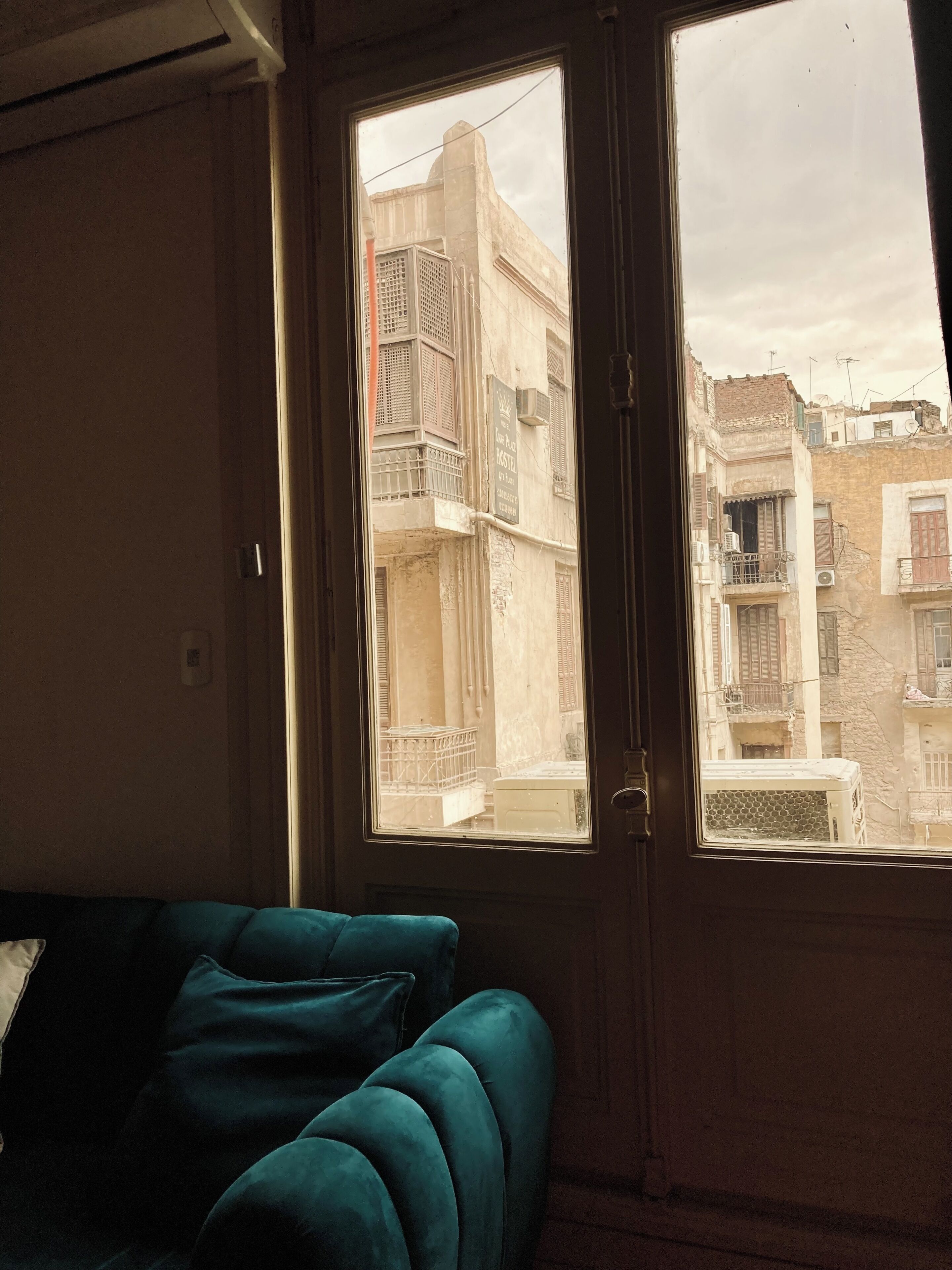 Una habitación con un sofá azul turquesa, un ventanal con vistas a edificios de varios pisos y una puerta.