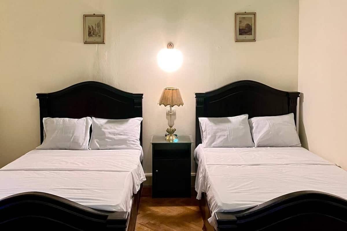 A room with two beds, a nightstand with a lamp, and a wall-mounted light fixture.