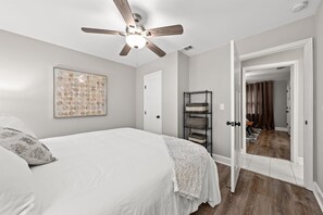 A bedroom with a bed, a ceiling fan, a wall art piece, a door, and a hallway leading to another room.