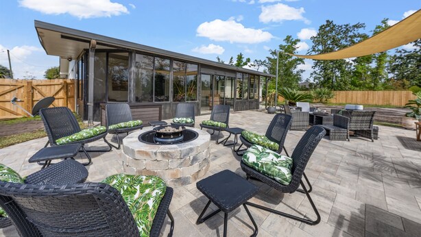 A modern outdoor patio with wicker furniture, a stone fire pit, and a glass-enclosed structure.
