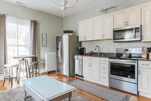 Una cocina moderna con armarios blancos, electrodomésticos de acero inoxidable y una zona de comedor con mesa de vidrio y taburetes de bar.