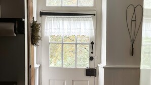 Um corredor com uma porta branca, uma janela com cortina de renda, piso de madeira e um candeeiro fixado na parede.