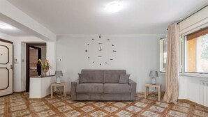 Un salon avec un canapé gris, deux tables d’appoint avec des lampes, une horloge murale et un sol à motifs.