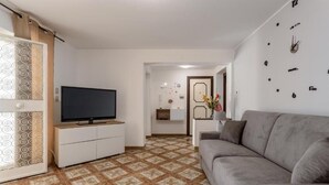 Un salon avec un canapé gris, un téléviseur à écran plat posé sur un meuble blanc, des carreaux de sol à motifs et une horloge murale.