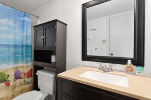 A bathroom with a white toilet, a beige sink, a dark cabinet, a shower curtain with a beach scene, and a large mirror.