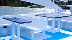 Un pont de bateau avec deux tables et des bancs blancs, un plancher bleu et une vue sur la mer et la plage en arrière-plan.