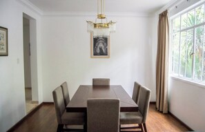 A dining room with a rectangular table, four chairs, a chandelier, a framed picture on the wall, and a window with curtains.