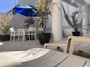 An outdoor seating area with white plastic chairs and tables, a blue umbrella, a hammock, and potted plants.