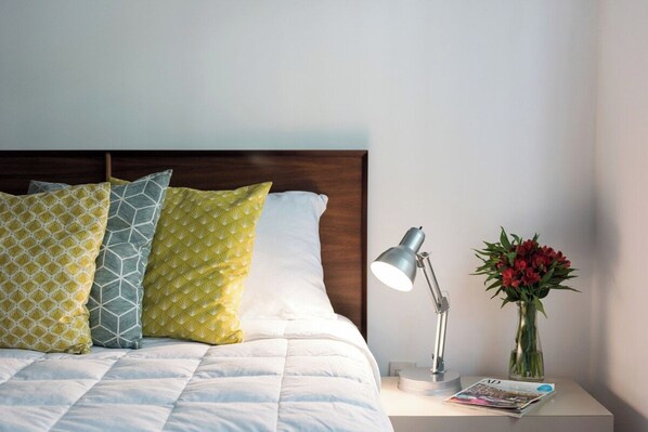A neatly made bed with patterned pillows, a bedside lamp, a magazine, and a vase with flowers on a nightstand.