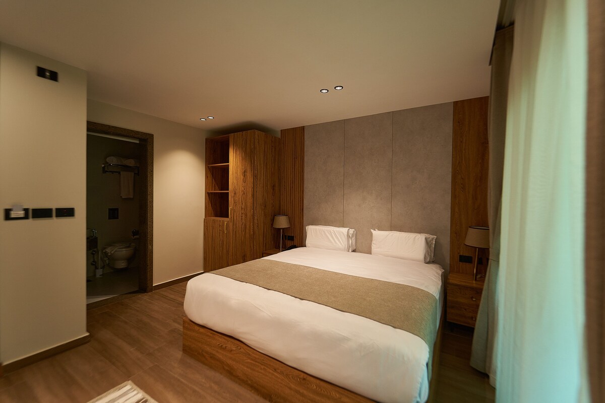 A modern hotel room with a large bed, wooden paneling, a built-in shelf, and a bathroom visible through an open door.