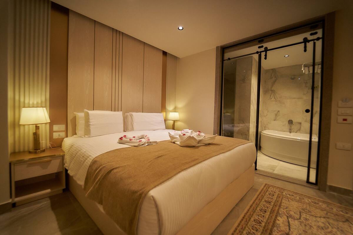 A hotel room with a large bed, bedside lamps, a glass shower enclosure, and a bathroom with a bathtub.