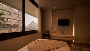 A hotel room with a bed, a window with a view of a pyramid, a flat-screen TV mounted on the wall, and a small table with a coffee maker. - Gold's Pyramids Hotel (Giza)