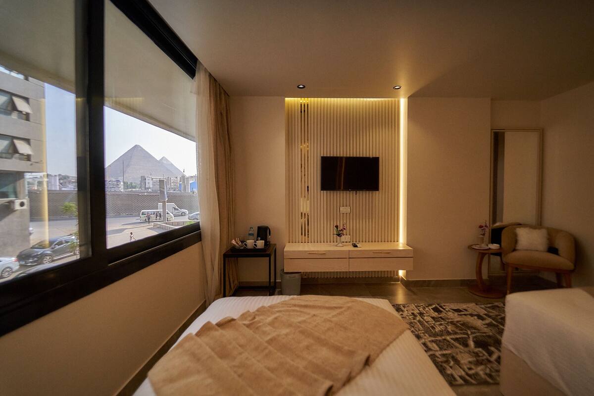 A hotel room with a bed, a bedside table with a lamp, a TV mounted on the wall, and a view of the pyramids through the window.