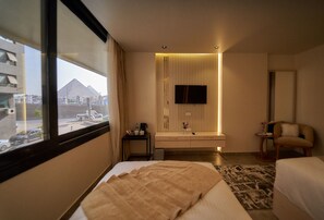 A hotel room with a bed, a bedside table with a lamp, a TV mounted on the wall, and a view of the pyramids through the window. - Gold's Pyramids Hotel (Giza)