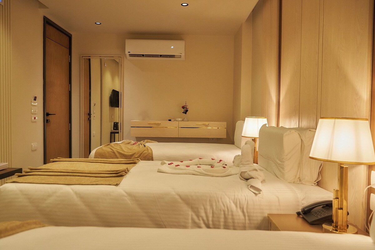 A hotel room with two beds, a bedside table with a lamp, a phone, and a small table with a vase.