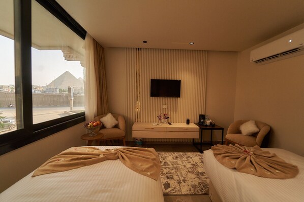 A hotel room with two beds, a desk, a chair, a TV, and a view of a pyramid. - Gold's Pyramids Hotel (Giza)