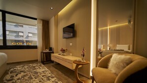 A hotel room with a bed, a sofa, a TV mounted on the wall, a small table with a vase, and a view of a building outside. - Gold's Pyramids Hotel (Giza)