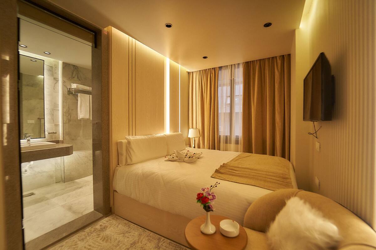 A hotel room with a bed, a bedside table with a vase, a TV mounted on the wall, and a bathroom with a glass shower enclosure.