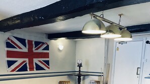 A pool table with a green felt surface, wooden legs, and a dark wood finish. There are two chairs with wooden seats and backs, and a table lamp with a metallic finish. A British flag is visible on the wall, and a door with a security camera is seen in the background. - Blue Bell (Kettering)